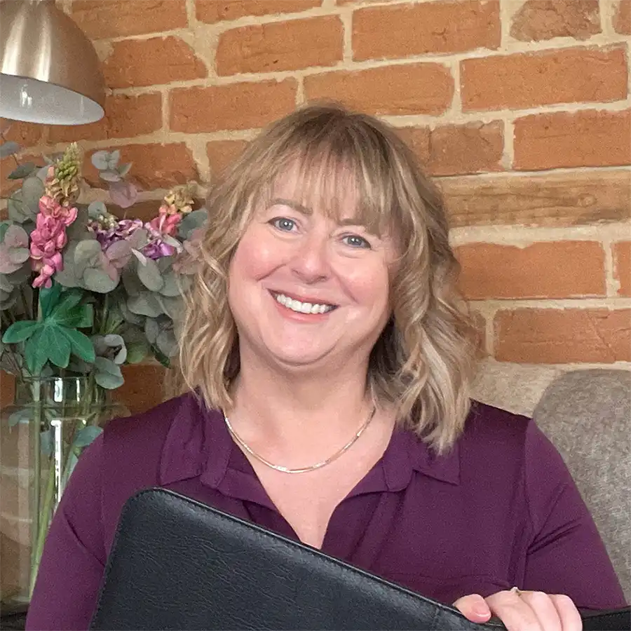 A smiling blonde woman with bangs wearing a purple collared shirt, holding a black folder in front of a brick wall background with a lamp and flowers.