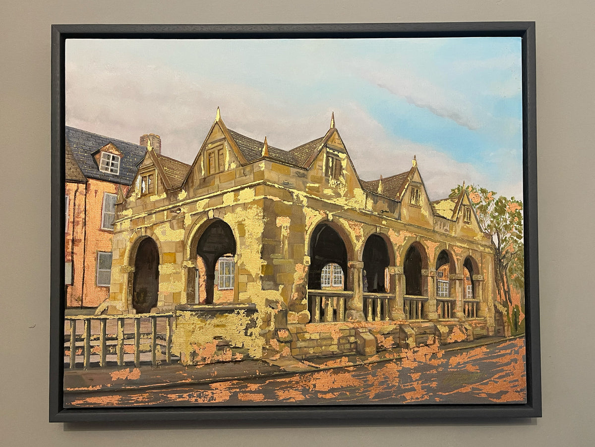 A framed painting of an old stone building with arches and gabled roofs, rendered in warm beige, brown, and orange tones under a blue sky, shown hanging on a light gray wall.